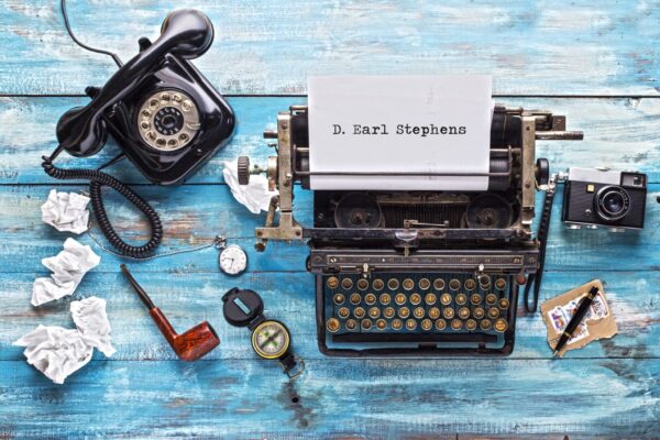 old typewriter with name typed on paper D. Earl Stephens. Blue colored distressed wood background, smoke pipe and rolled up paper